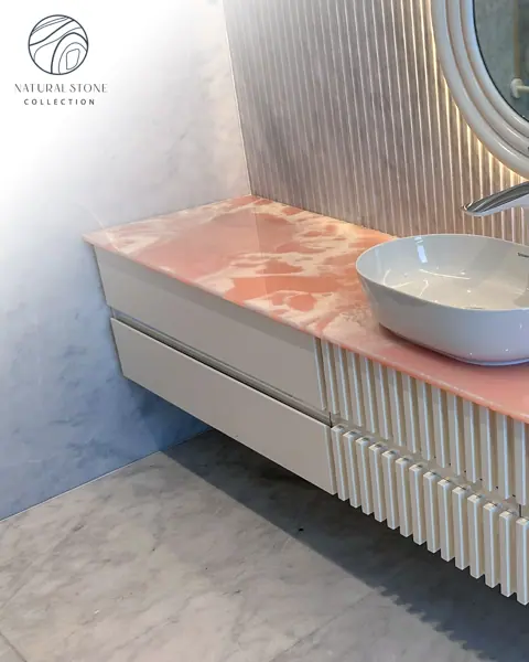 Rose coloured veined stone is used for the vanity and its surrounding surfaces, alongside vertical timber detailing and warm concealed lighting.
