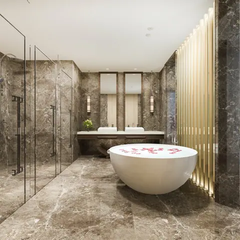 Rich brown veined stone is used for the bathroom walls and floor, alongside a freestanding white bath and soft lighting.