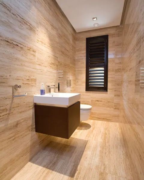 Warm beige natural stone is used for the vanity and its surrounding surfaces, alongside dark taps and a sculptural basin.