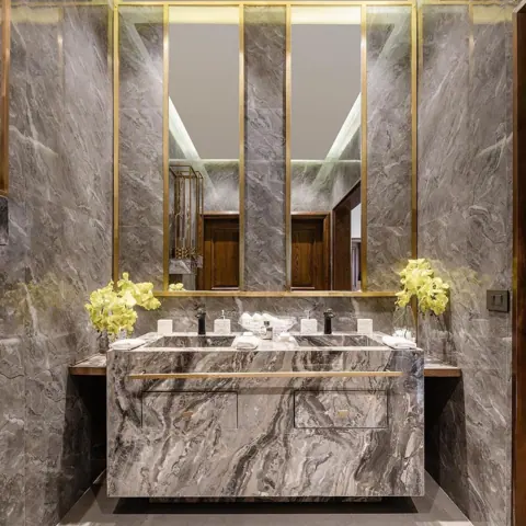 Grey veined stone is used for the vanity and its surrounding surfaces, alongside warm metallic details and carefully placed lighting. Golden mirror frames add a vertical rhythm behind the basin.