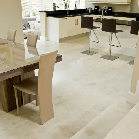Soft cream stone is used for the floor throughout the room, alongside white dining chairs and a light interior palette. The stone continues from the dining area toward the adjoining kitchen.