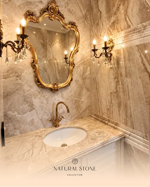 Warm veined stone surrounds a white vanity and oval basin, while a gilded mirror and decorative wall lights complete the classical setting.