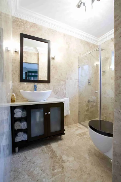 Warm beige stone covers the bathroom walls and floor, paired with a dark vanity, a framed mirror and a glass shower enclosure.